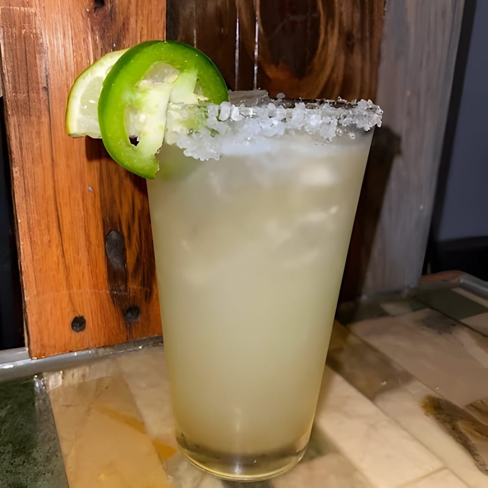 A margarita with a salted rim, garnished with lime and jalapeño slices, against a wooden background.
