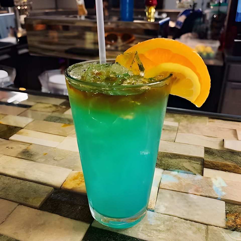 A vibrant blue cocktail with crushed ice, a white straw, and orange slices on a tiled bar.