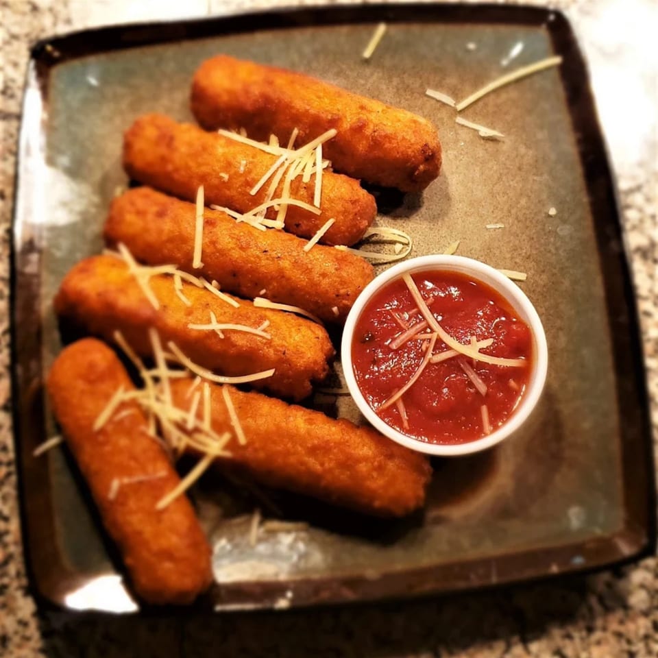 Golden-brown mozzarella sticks with shredded cheese and a side of marinara sauce on a dark plate.